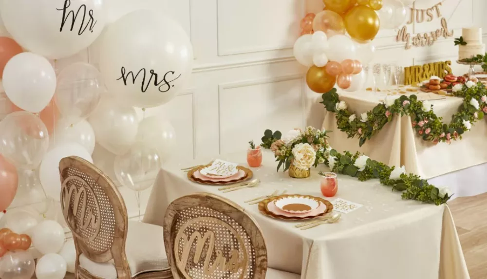 Two tables in a white room decorated fpr a wedding with white, gold and pink balloons