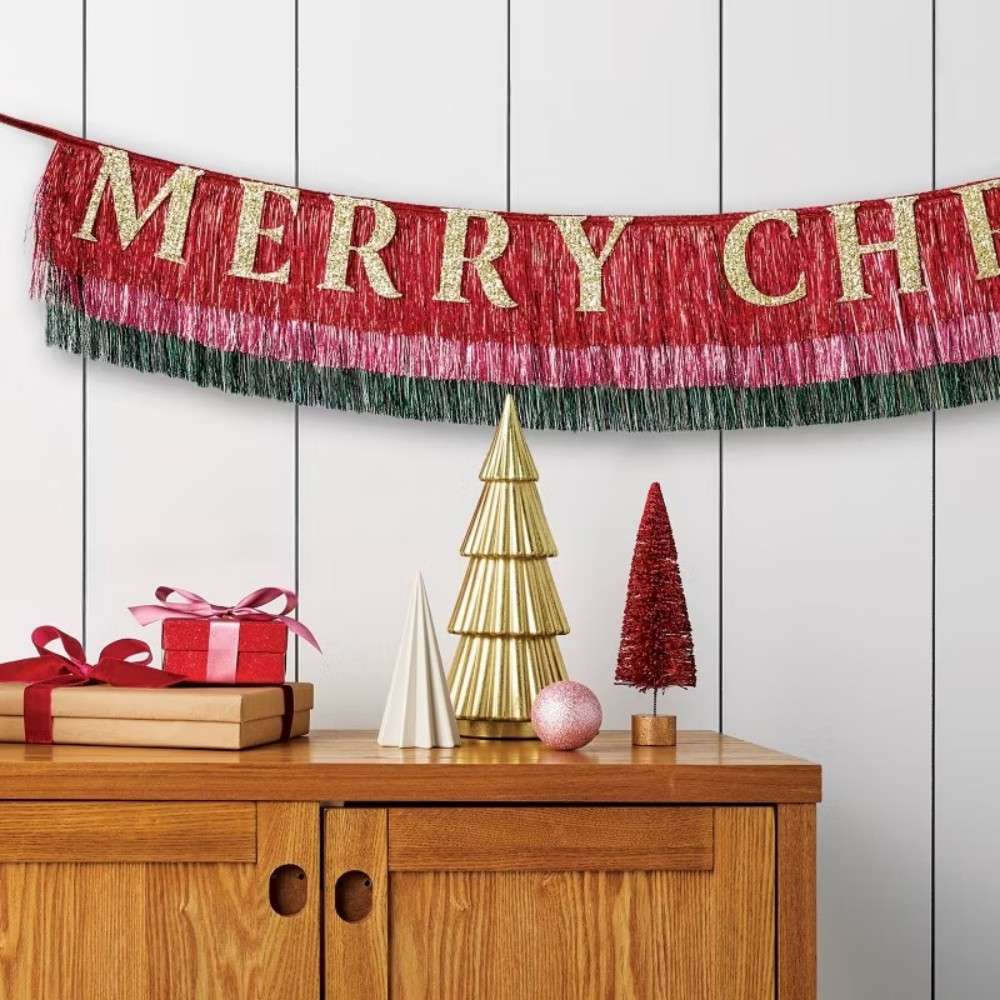 A red, pink, and green colored garland with gold colored letters spelling _MERRY CH_ hangs above a wooden cabinet decorated with small cone-shaped trees, a pink ornament, and wrapped gifts.