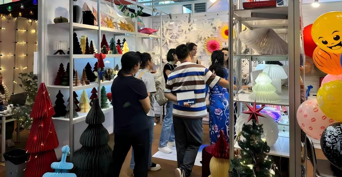 Trade show booth displaying Sunbeauty Factory's festive paper decorations (Christmas trees, stars, snowflakes) with visitors browsing.