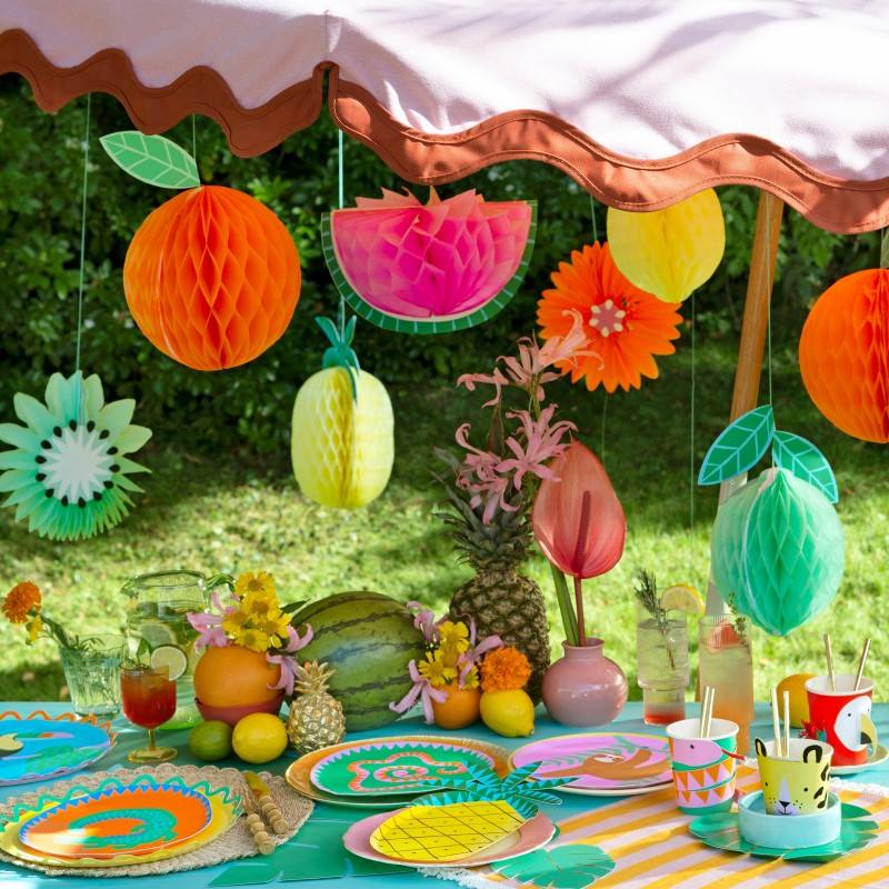 Summer outdoor party table setup with hanging tropical fruit honeycomb decorations, colorful plates, and drinks