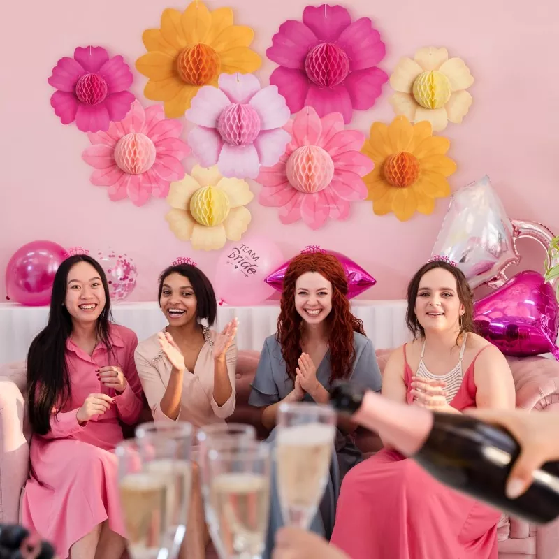 Bachelorette party scene with women celebrating, large honeycomb paper flower backdrop on pink wall and bride balloons