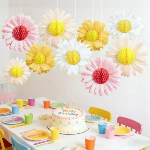 Hanging pink white yellow daisy honeycomb paper flowers suspended over indoor birthday table with birthday cake and bright party cups