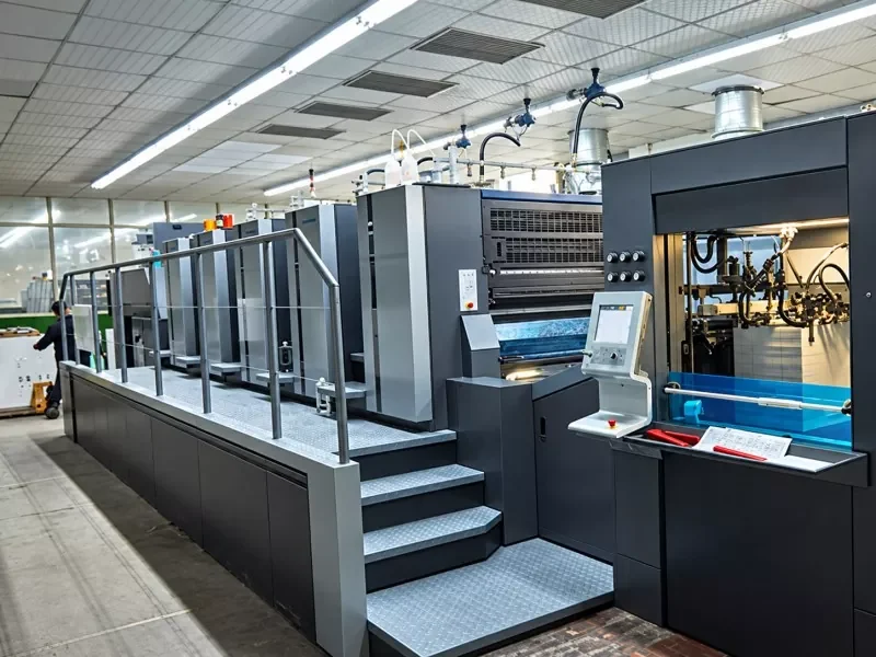 Large‑format offset printing press inside bright manufacturing plant, industrial print production machinery with control panel and work platform